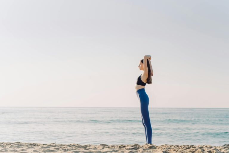 A woman performing yoga on a serene beach with a clear blue sky, emphasizing relaxation and wellness.