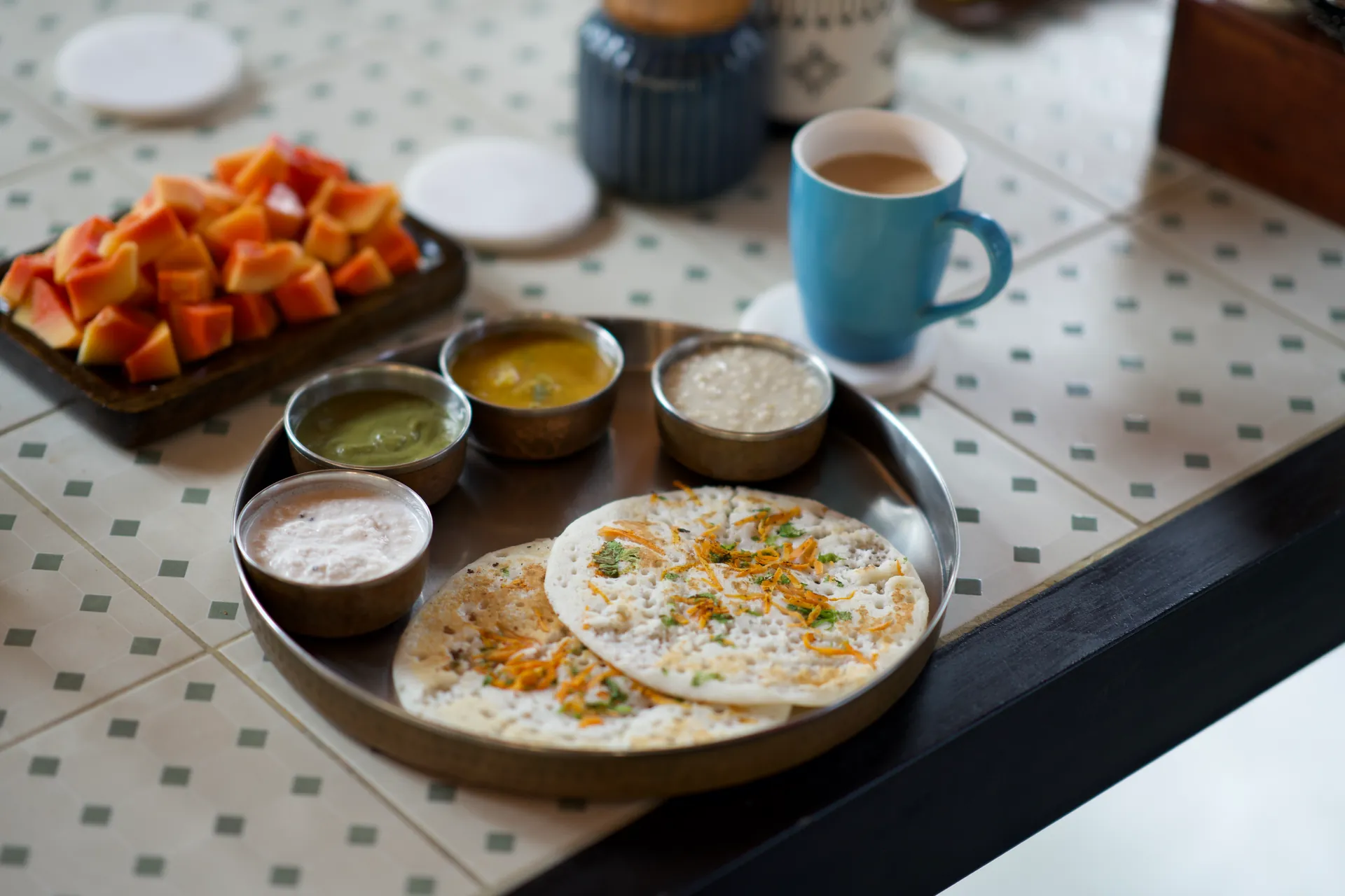 Steel plate of uttapam and chutneys next to papaya and a blue mug of chai