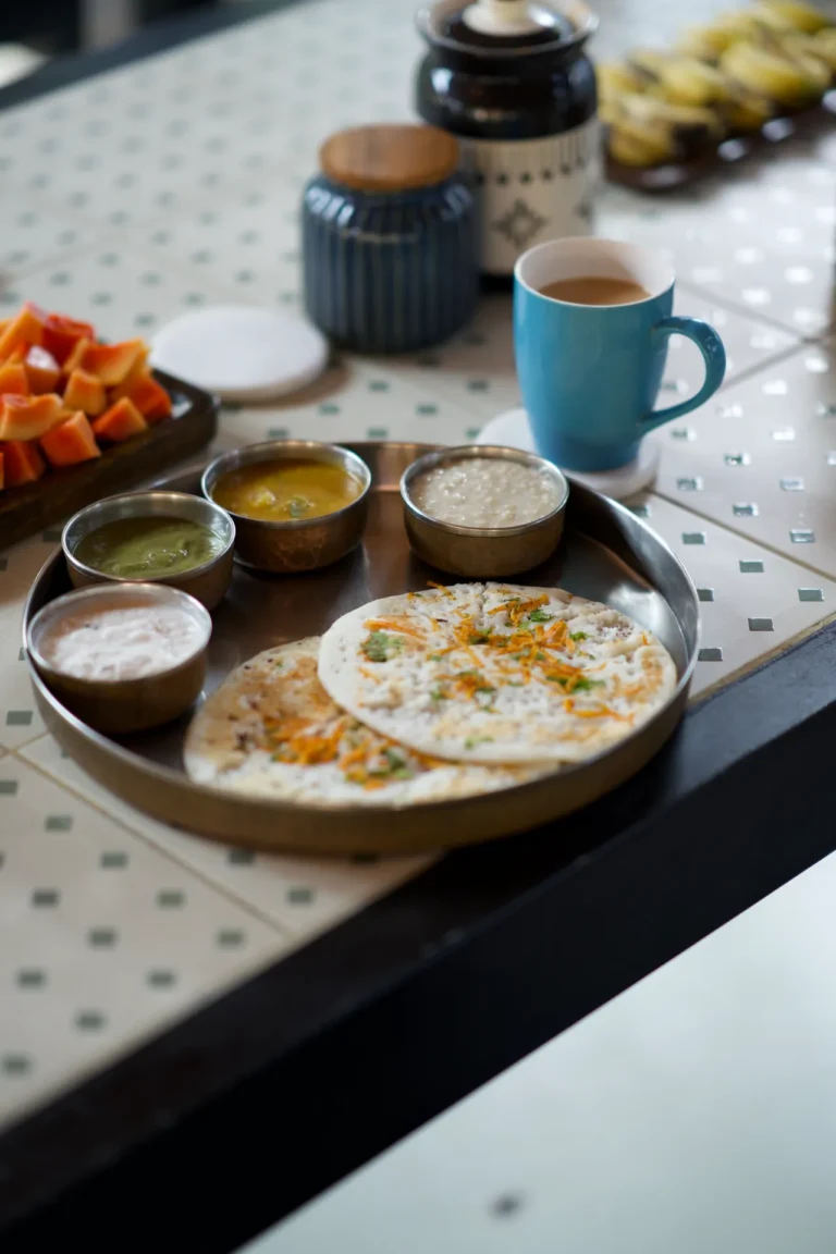 Two uttapam with small bowls of chutney, a plate of papaya and a cup of chai on a tiled table