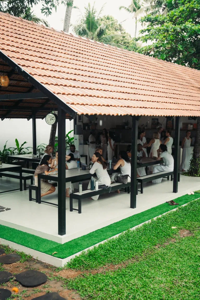 Students in white eating and chatting at long tables under the tiled roof of the dining pavilion
