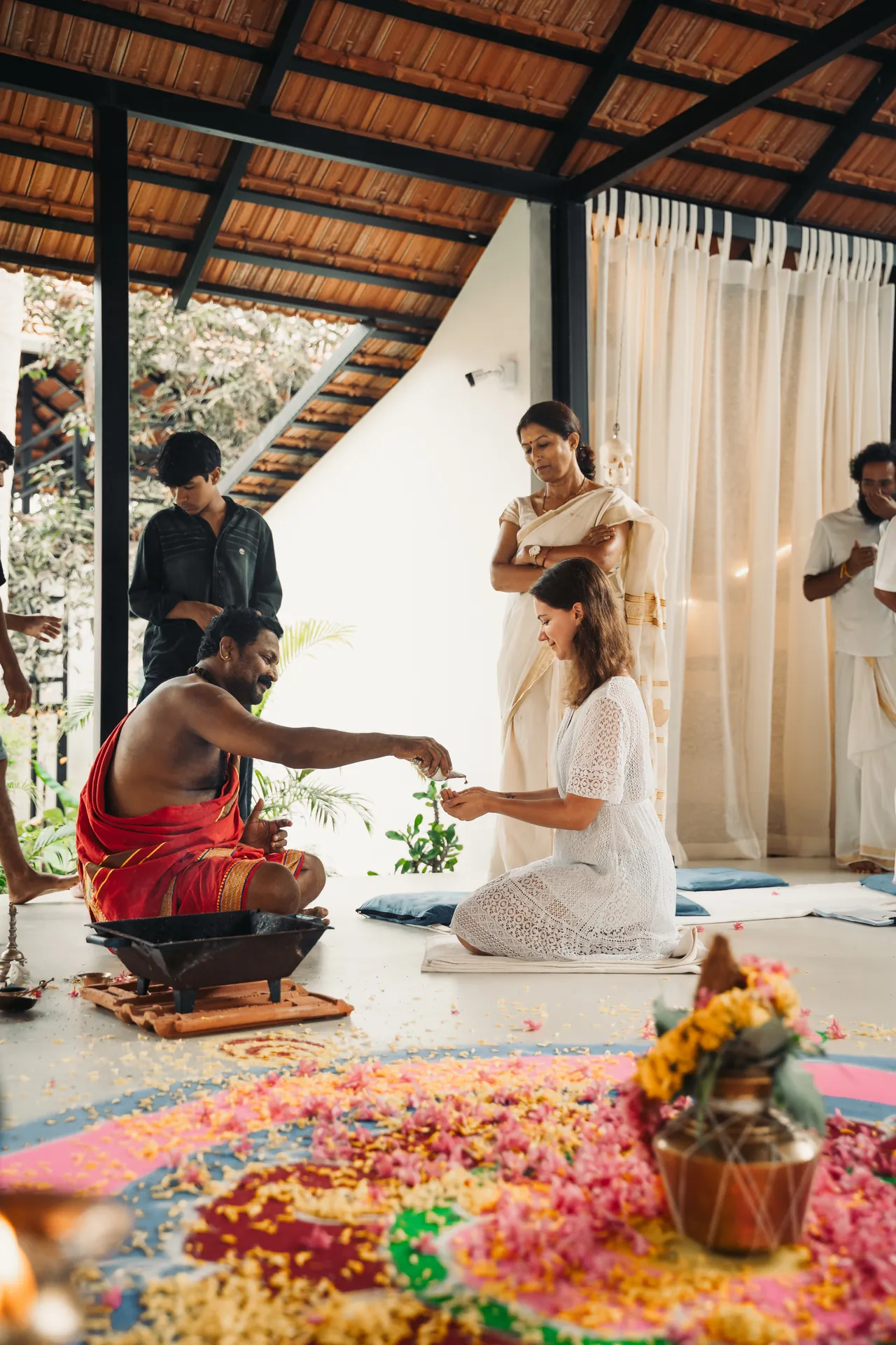 Student in white kneeling to receive an offering from a man in a red dhoti beside a flower rangoli