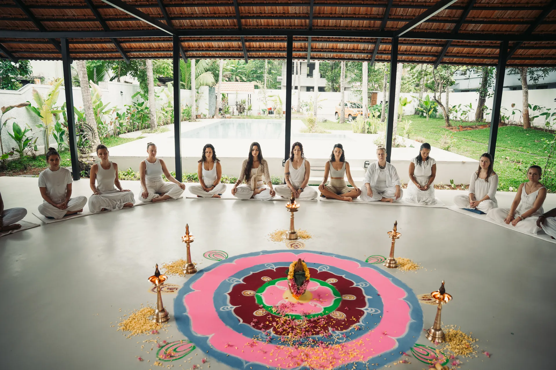 Students in white sitting in a circle around a flower rangoli and brass lamps in the open shala