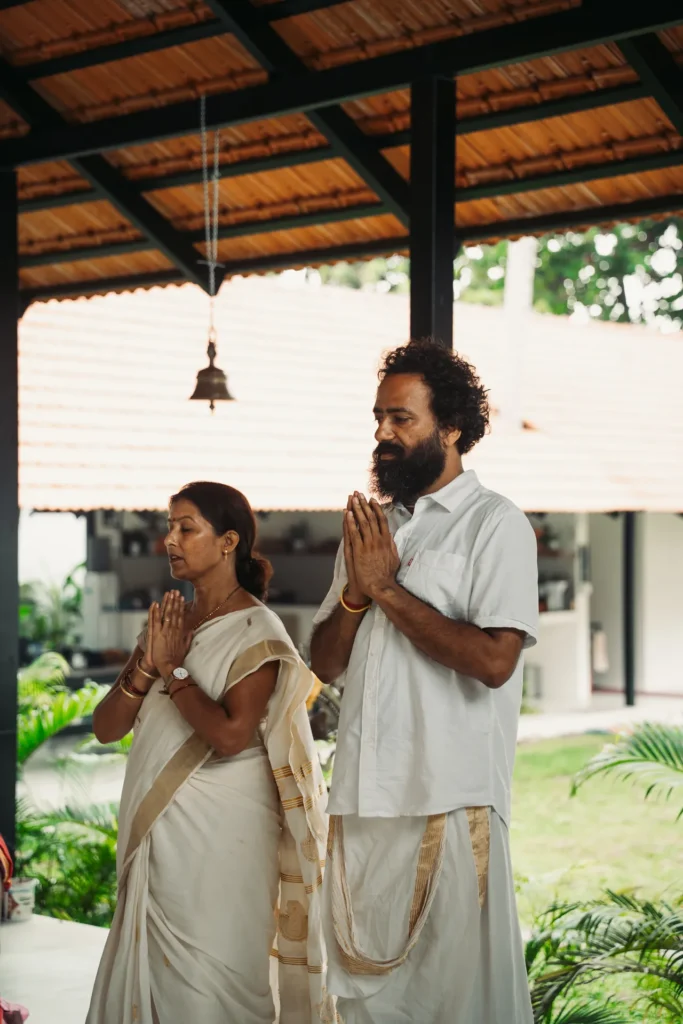 Woman in a Kerala sari and a bearded man standing with palms together under the shala roof